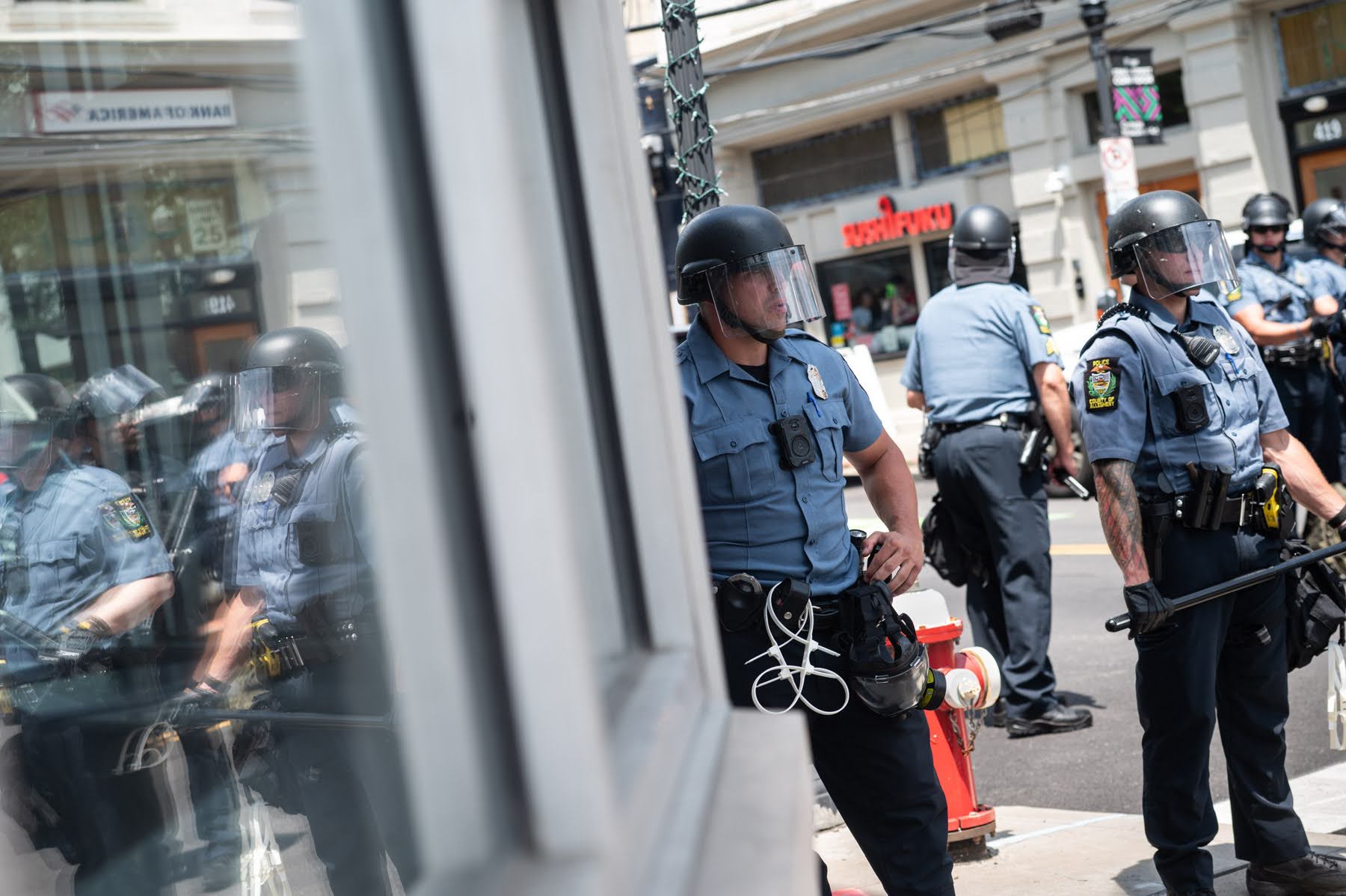 Four police officers in riot gear stand on a city sidewalk, with their reflections visible in a nearby glass window.