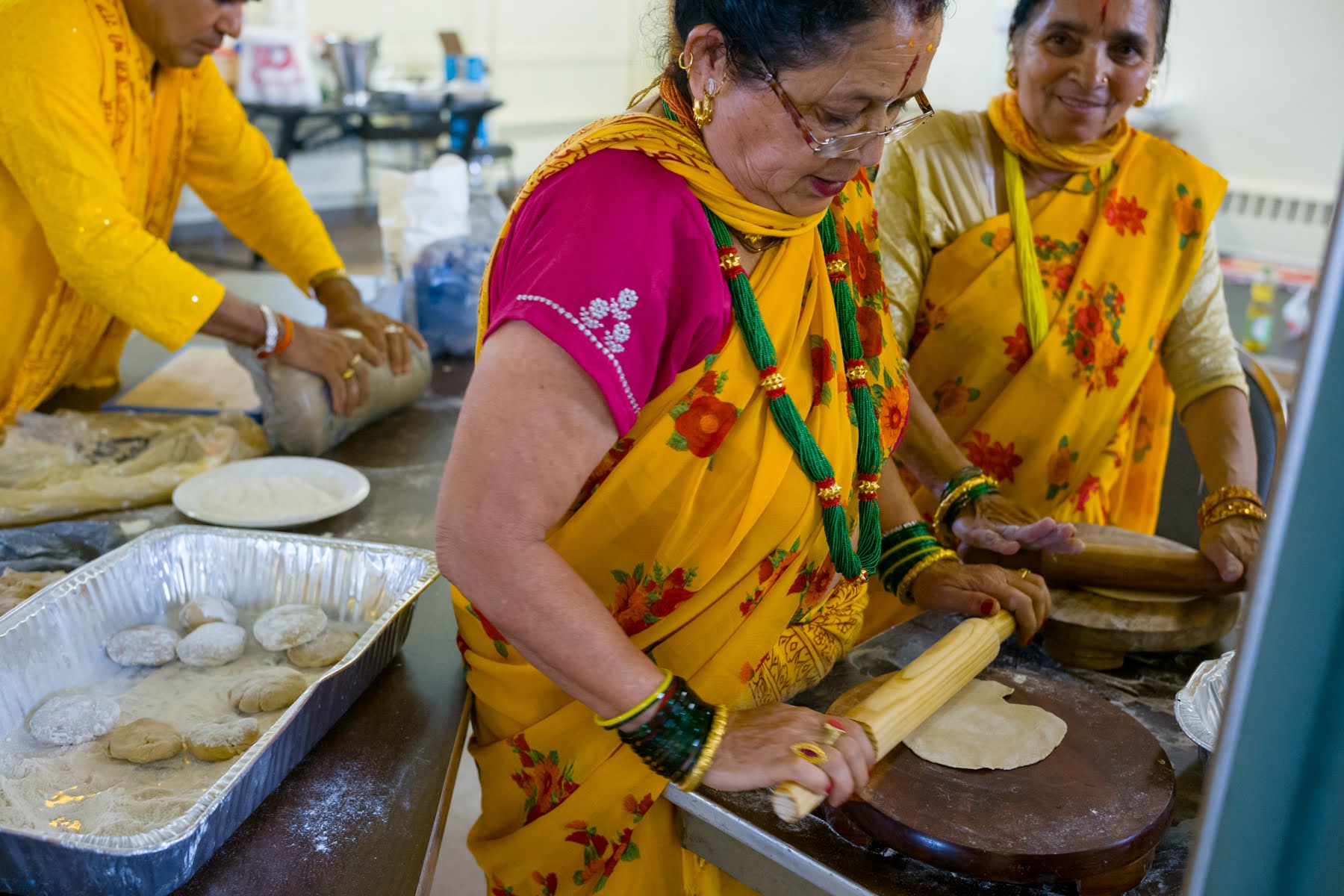 Three women in colorful traditional attire roll out dough and prepare flatbreads on a table covered with flour and baking trays.