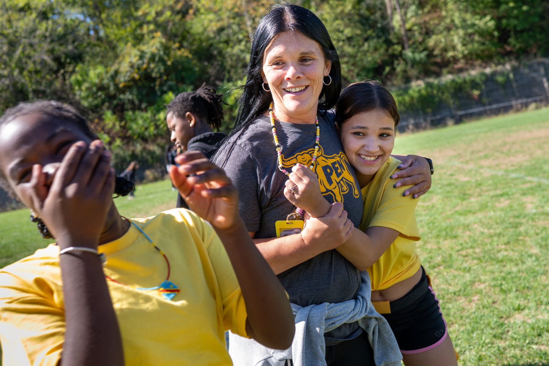 A woman stands smiling on a grassy field with two children, one hugging her and another laughing, on a sunny day with trees in the background.