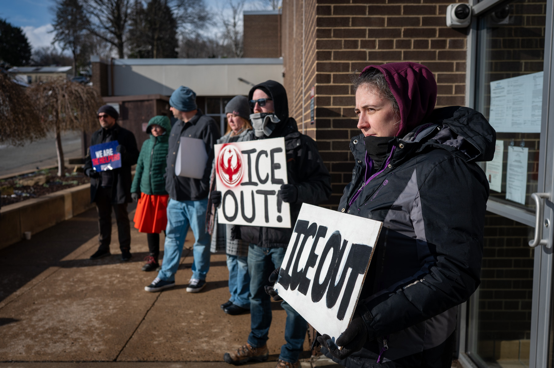 A group of people stand outside a building in winter clothing, holding signs that read "ICE OUT!" during a protest.