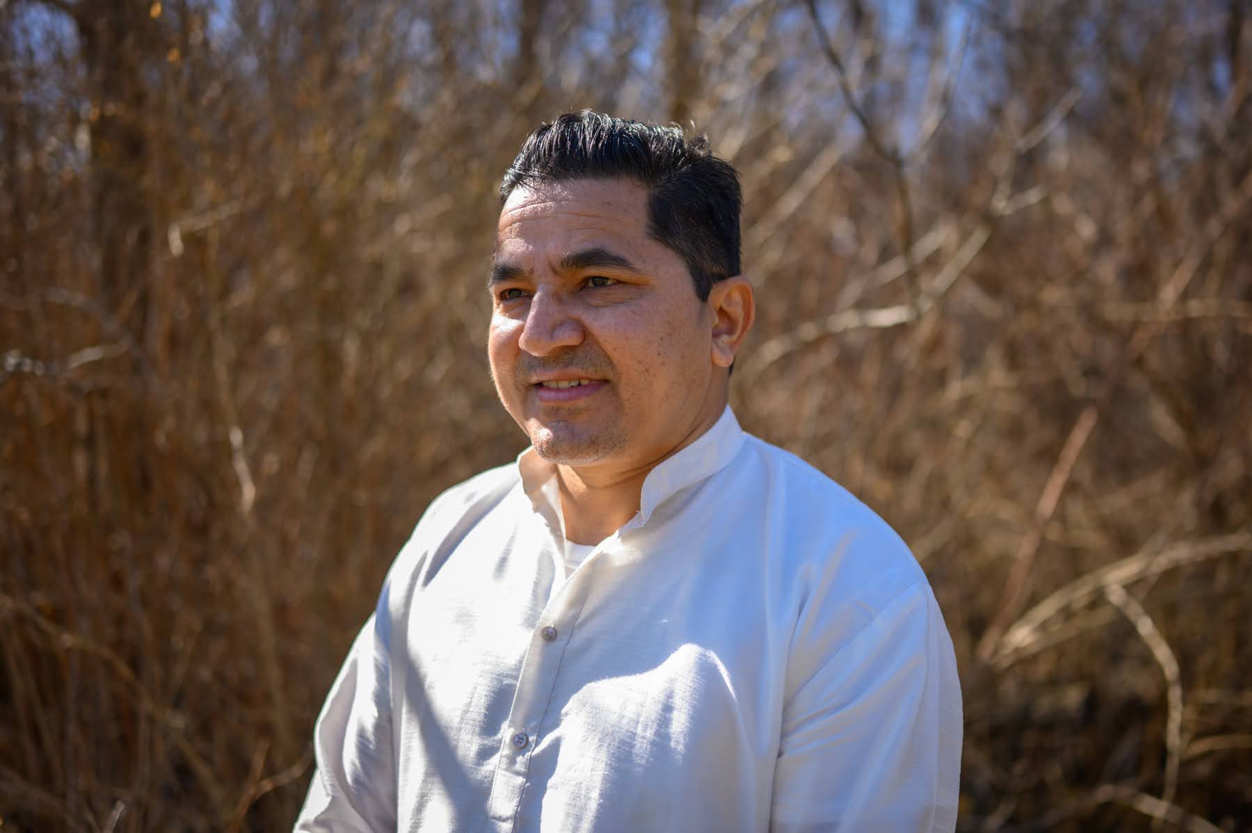 A man wearing a white shirt stands outdoors in front of dry, brown vegetation under bright sunlight.