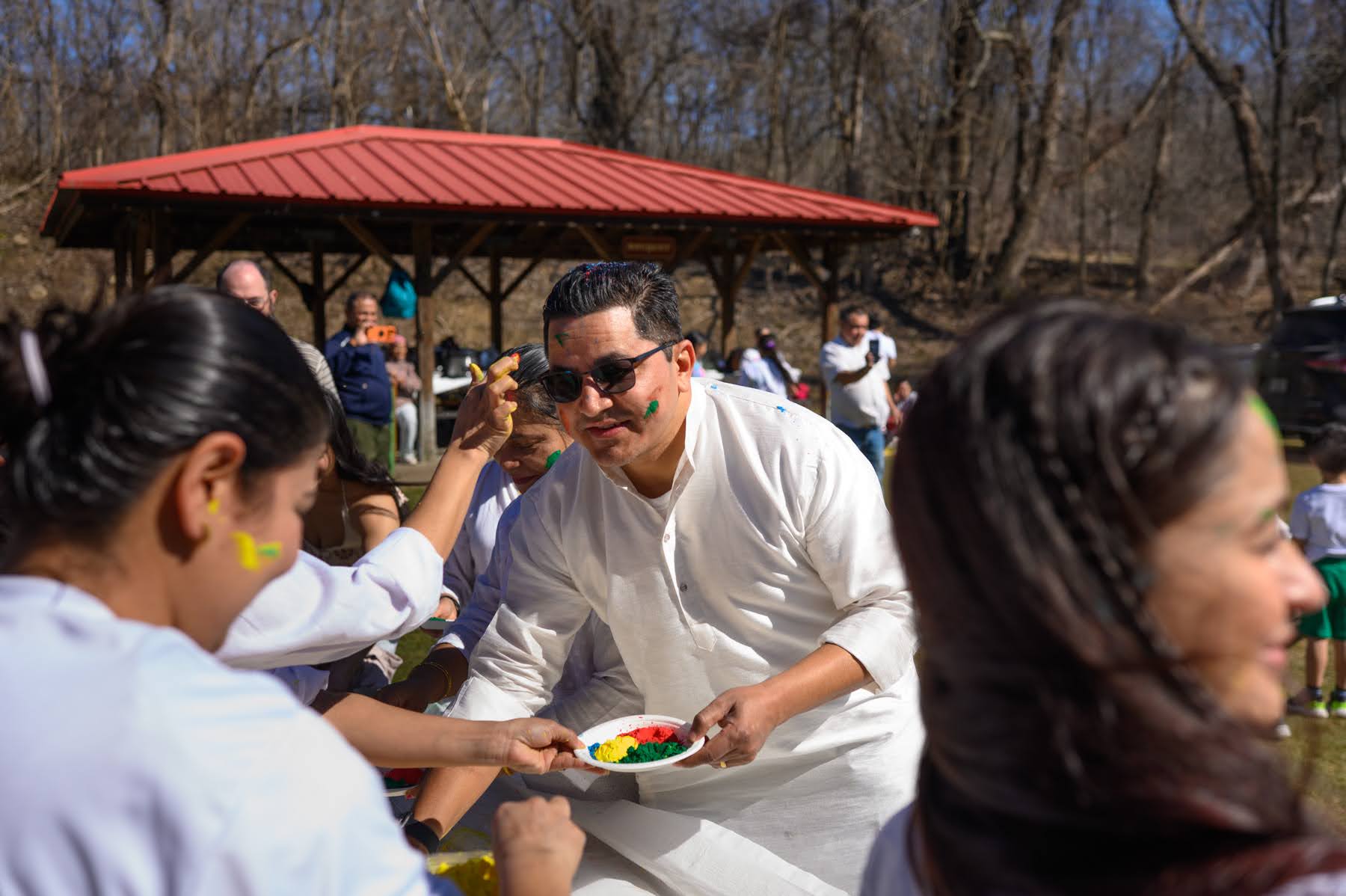People wearing white clothes celebrate Holi outdoors, applying colored powder to each other. A man in sunglasses holds a plate of colored powder. A red pavilion and trees are in the background.