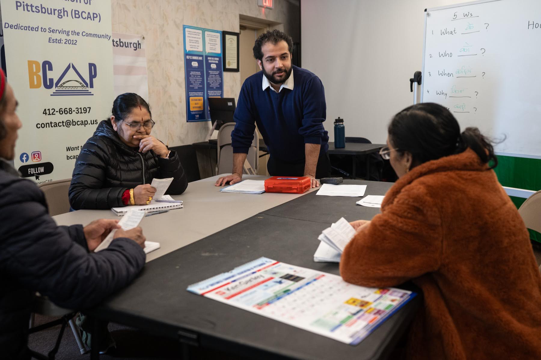 A man stands and talks to three seated adults in a classroom setting with papers, a calendar, and a whiteboard visible.