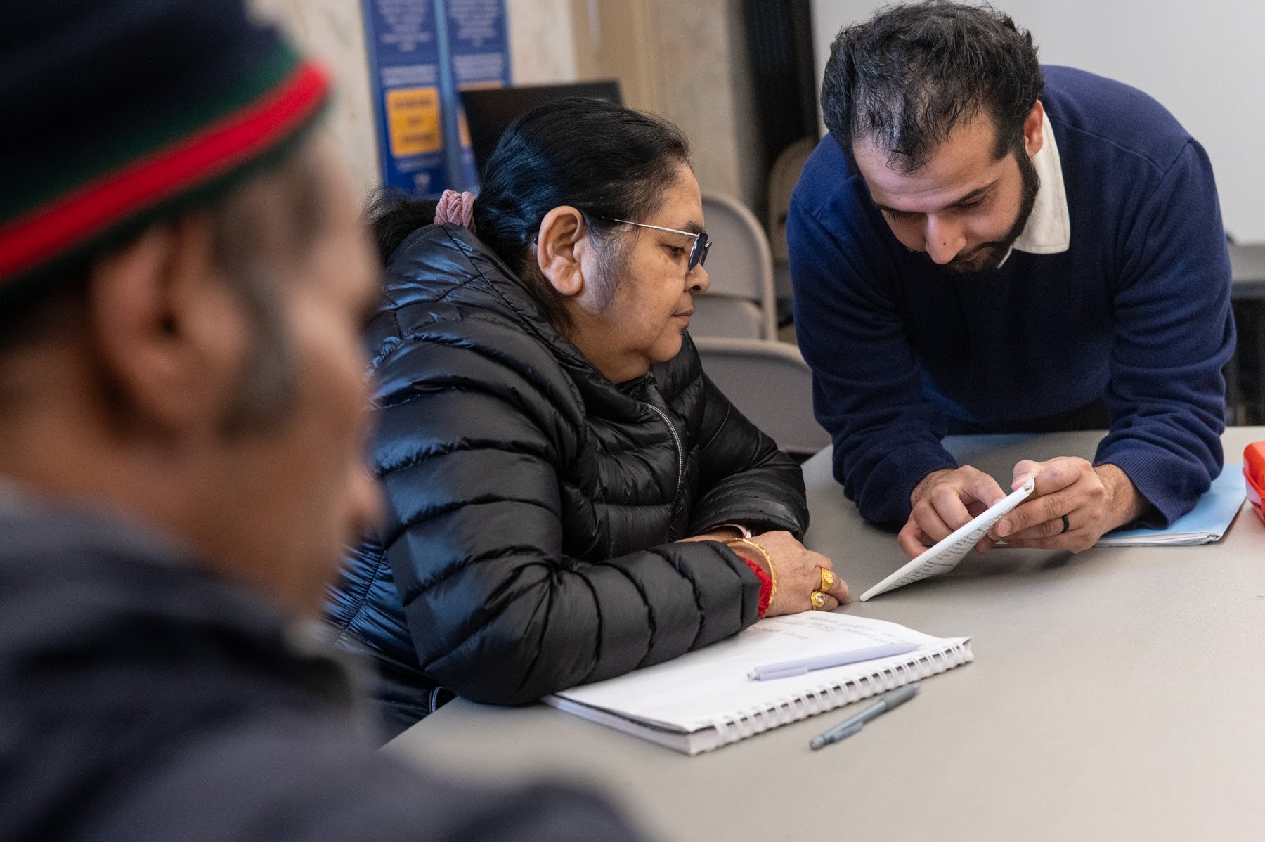 A man leans over to explain something on a sheet of paper to a seated woman during a meeting, while another person sits in the foreground.