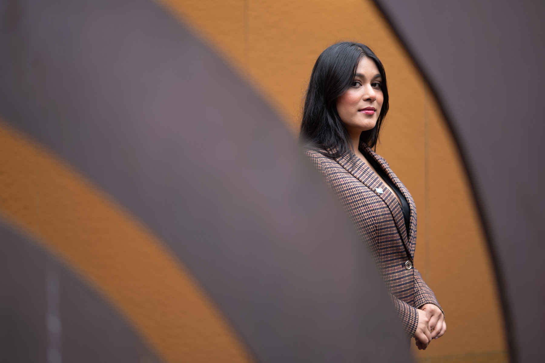 A woman in a checked blazer stands in front of an orange wall, framed by curved, blurred metal structures in the foreground.