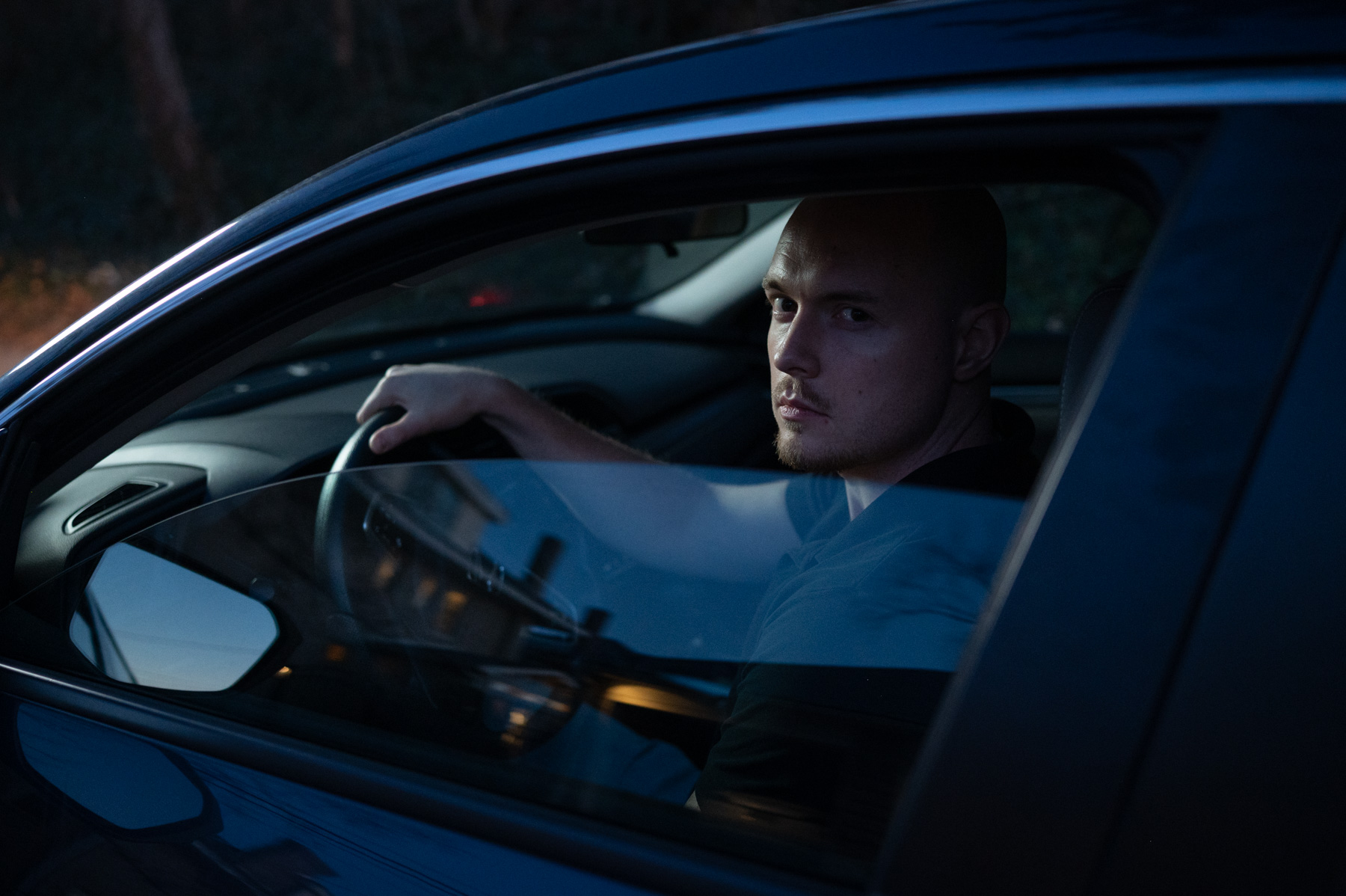 A man with a shaved head sits in the driver's seat of a car, looking out the open window during dusk.