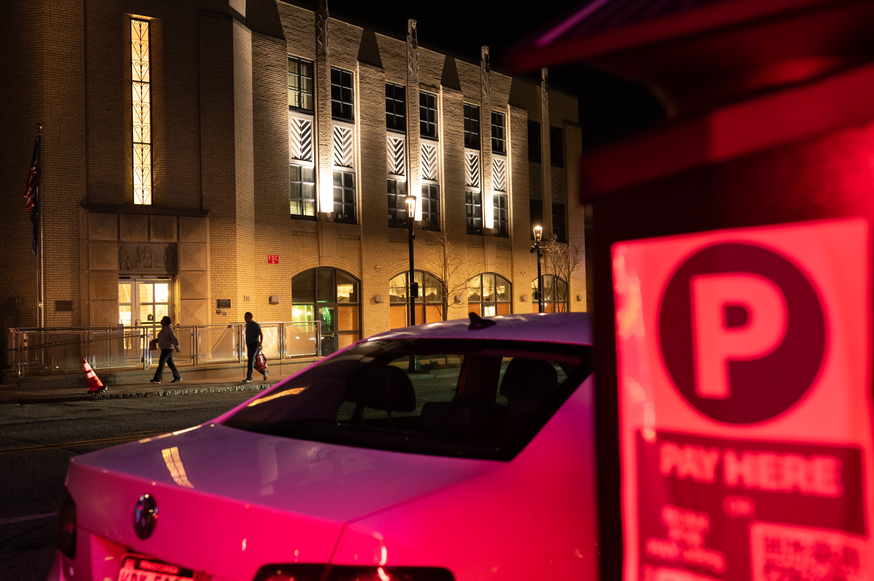 A white car is parked near a pay station on a city street at night, with a lit building and pedestrians visible in the background.