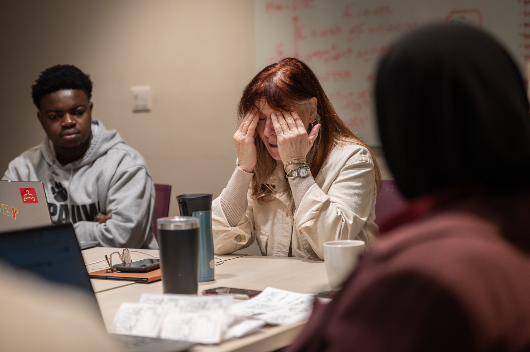A red-haired woman rubs her forehead while seated at a table with several young people in a conference room.