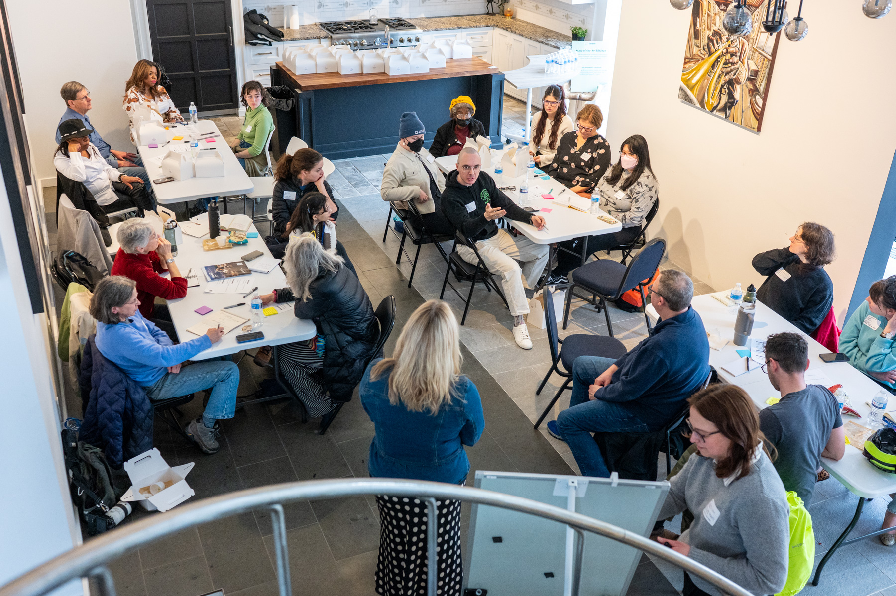 A group of people sit and stand around tables in a bright room, engaged in discussion or a workshop, with notepads and food containers visible.