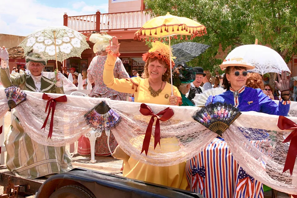People in vintage-style clothing wave while riding a parade float decorated with lace and fans
