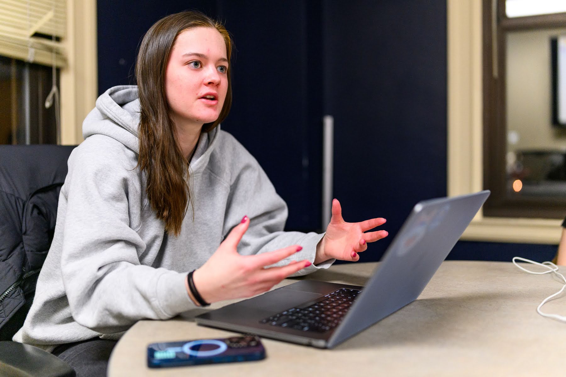 A woman in a gray hoodie sits at a table, gesturing with her hands while talking, with an open laptop and a smartphone in front of her.