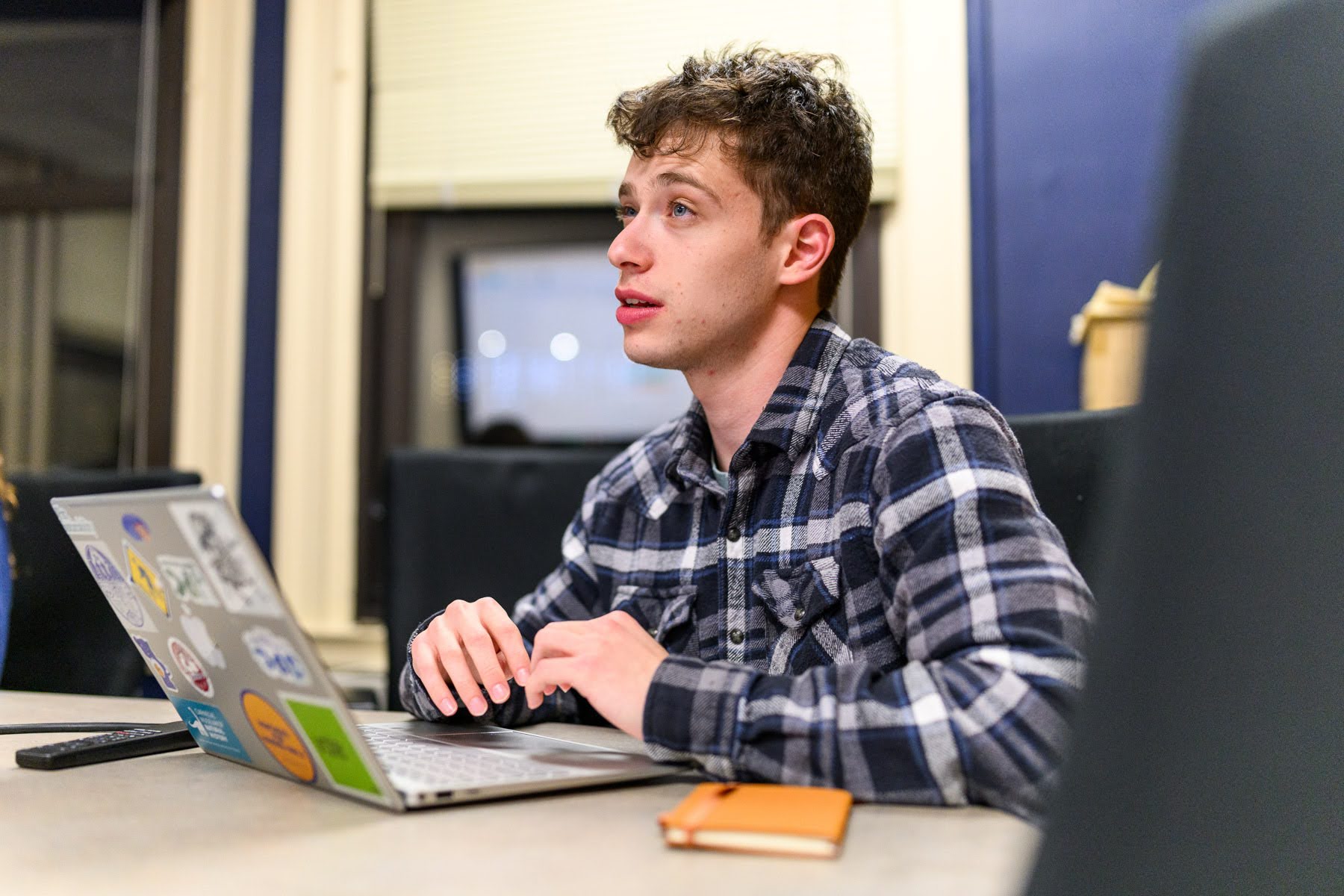 A young man in a plaid shirt sits at a table, using a sticker-covered laptop, with a notebook and pen nearby. He appears to be speaking or listening attentively.
