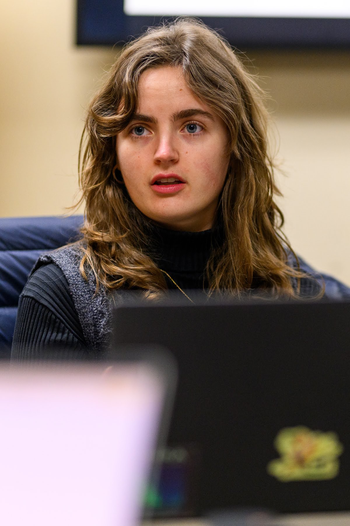 A young woman with wavy brown hair and a black turtleneck sits indoors, looking slightly to the side. A blurred laptop appears in the foreground.