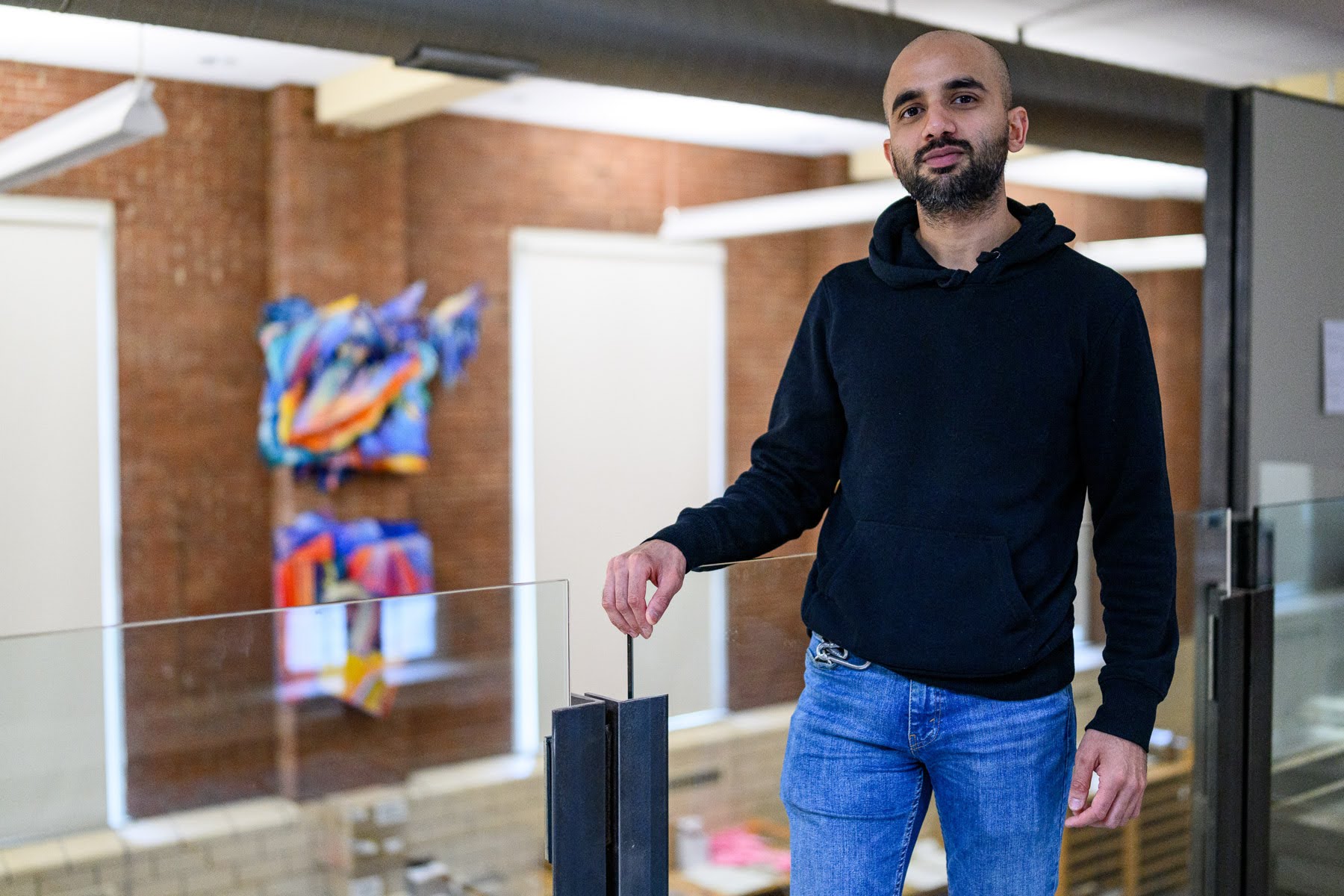A person in a black hoodie and blue jeans stands indoors by a glass railing, with brick walls and colorful abstract art in the background.