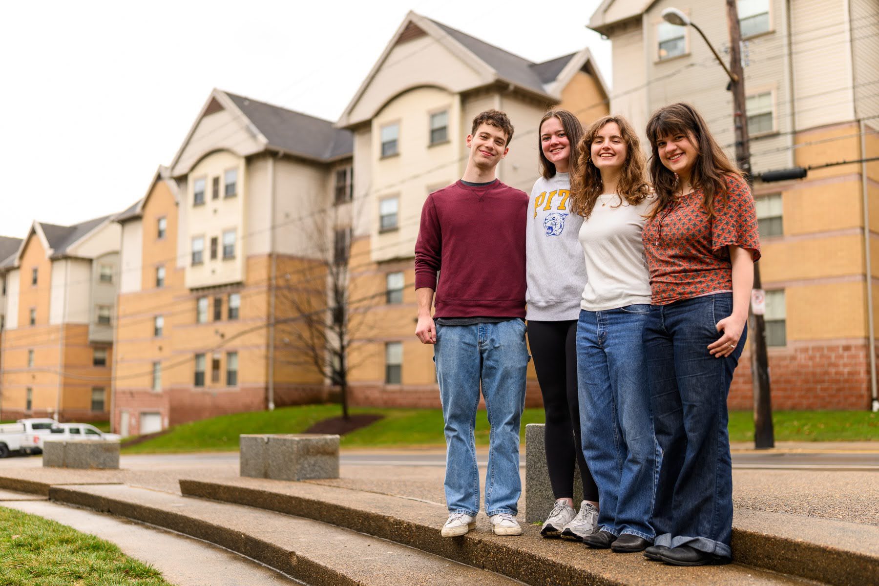 Four young adults stand together outdoors, smiling in front of apartment buildings on a cloudy day.