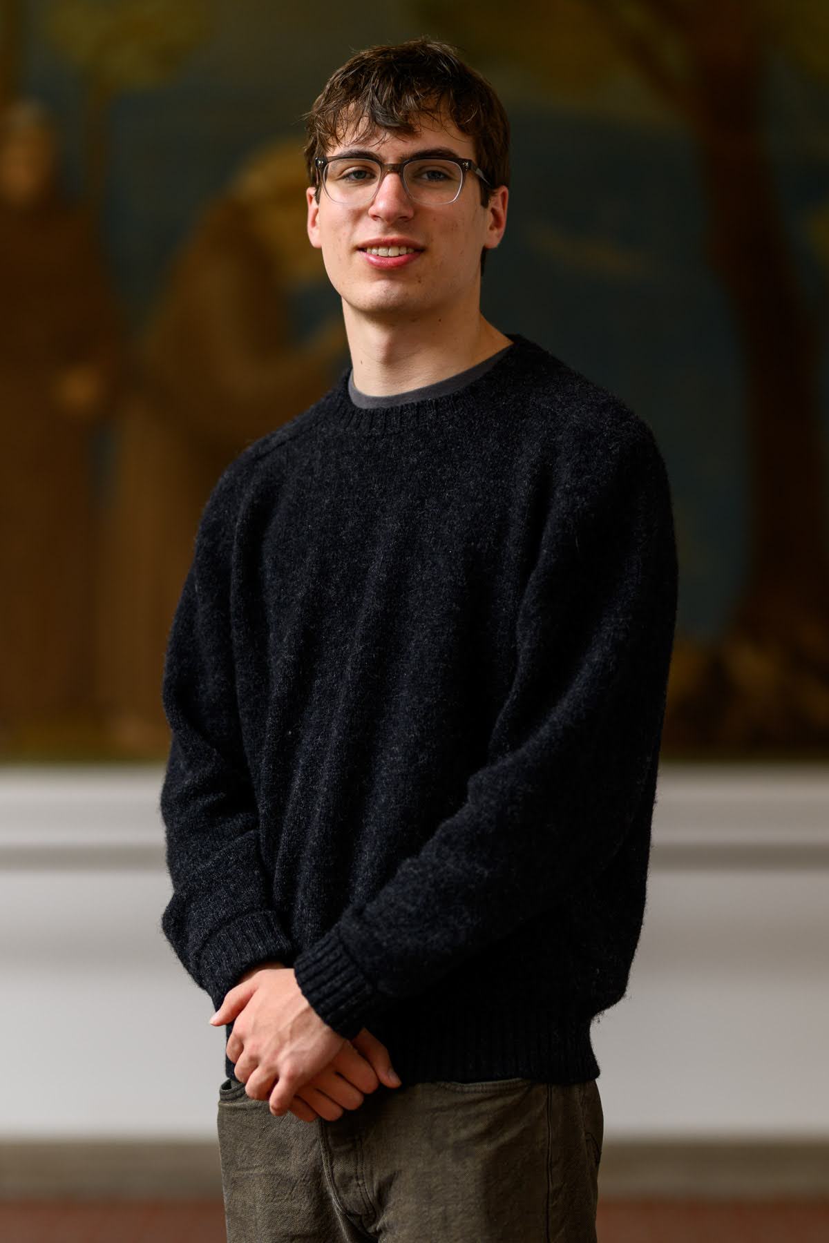 A young person wearing glasses, a black sweater, and dark pants stands indoors with hands clasped, looking at the camera. A blurred painting is visible in the background.
