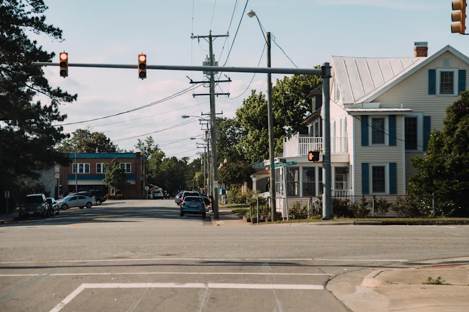 A quiet street intersection with a car waiting at a traffic light near houses and trees