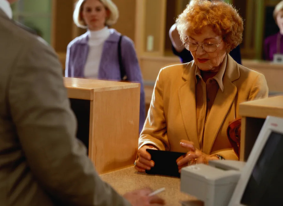 Elderly woman at bank counter holding a wallet, likely speaking to a bank teller. Other customers wait in line