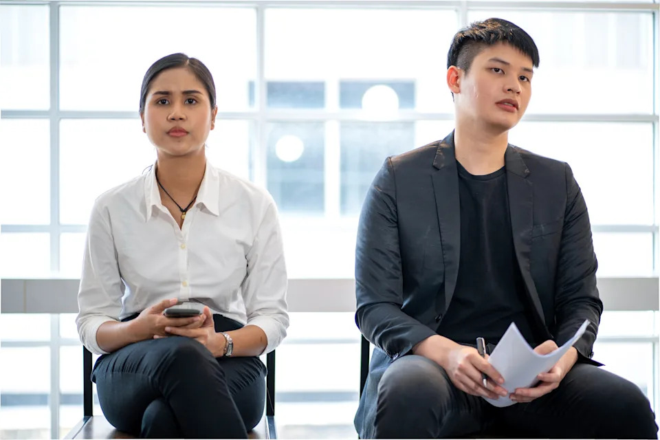 Two people sit in a waiting area. The person on the left wears a white shirt and holds a phone. The person on the right wears a black jacket and holds papers