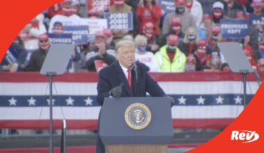 Donald Trump stands at a podium at a rally, with a crowd behind him.