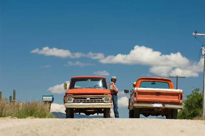 Two vintage trucks parked on a rural road, with a person in a hat standing between them, engaging in conversation. Sparse clouds are visible in the sky