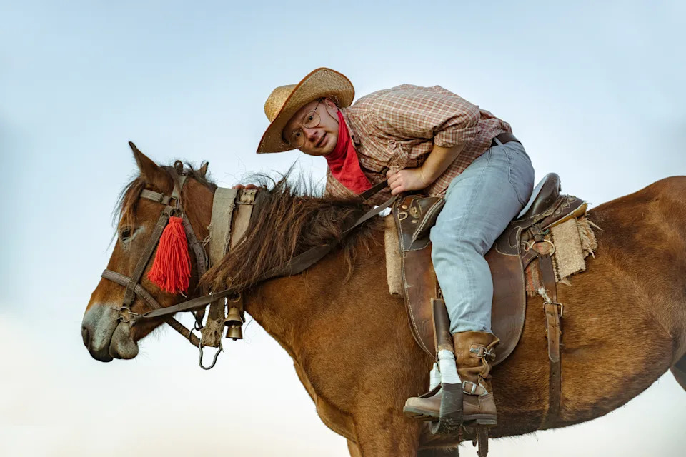 Person wearing a cowboy hat and plaid shirt playfully leans on a horse's neck, outdoors