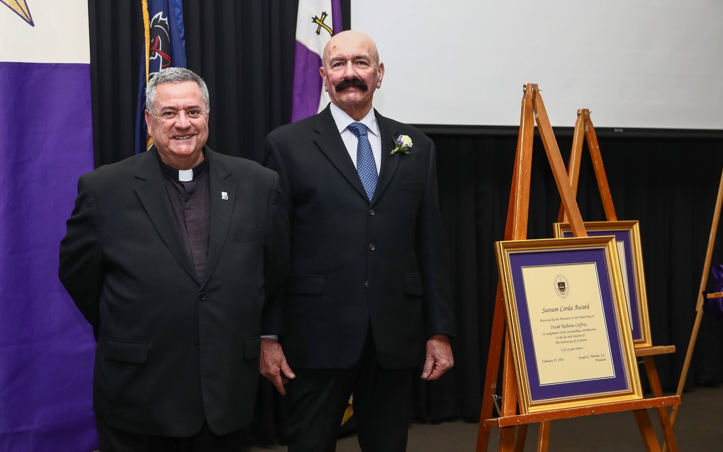 Two individuals in formal attire stand beside an easel displaying a framed certificate.