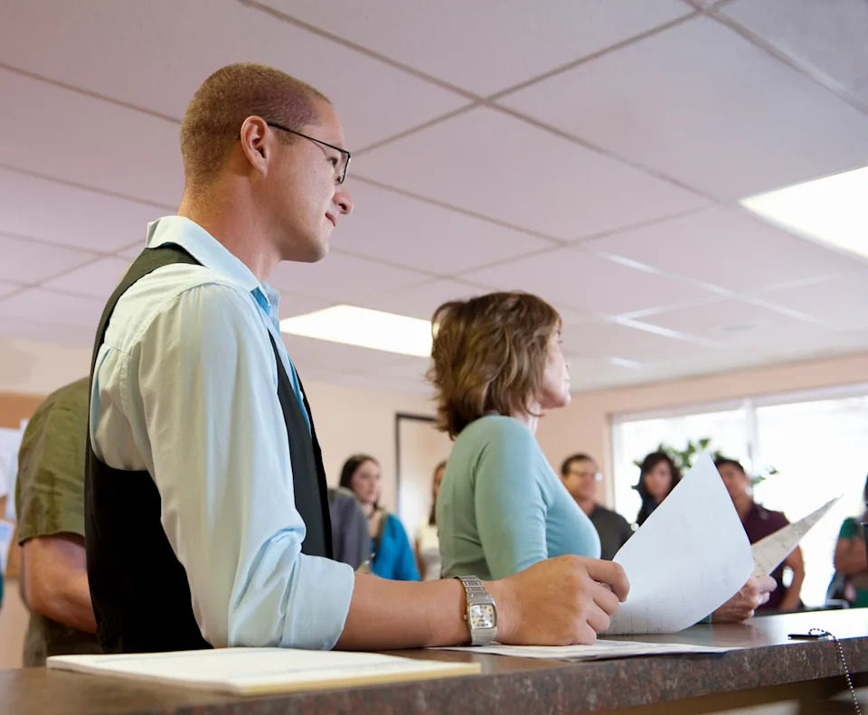 A man in a vest and a woman hold papers, speaking to a gathered group in an indoor setting