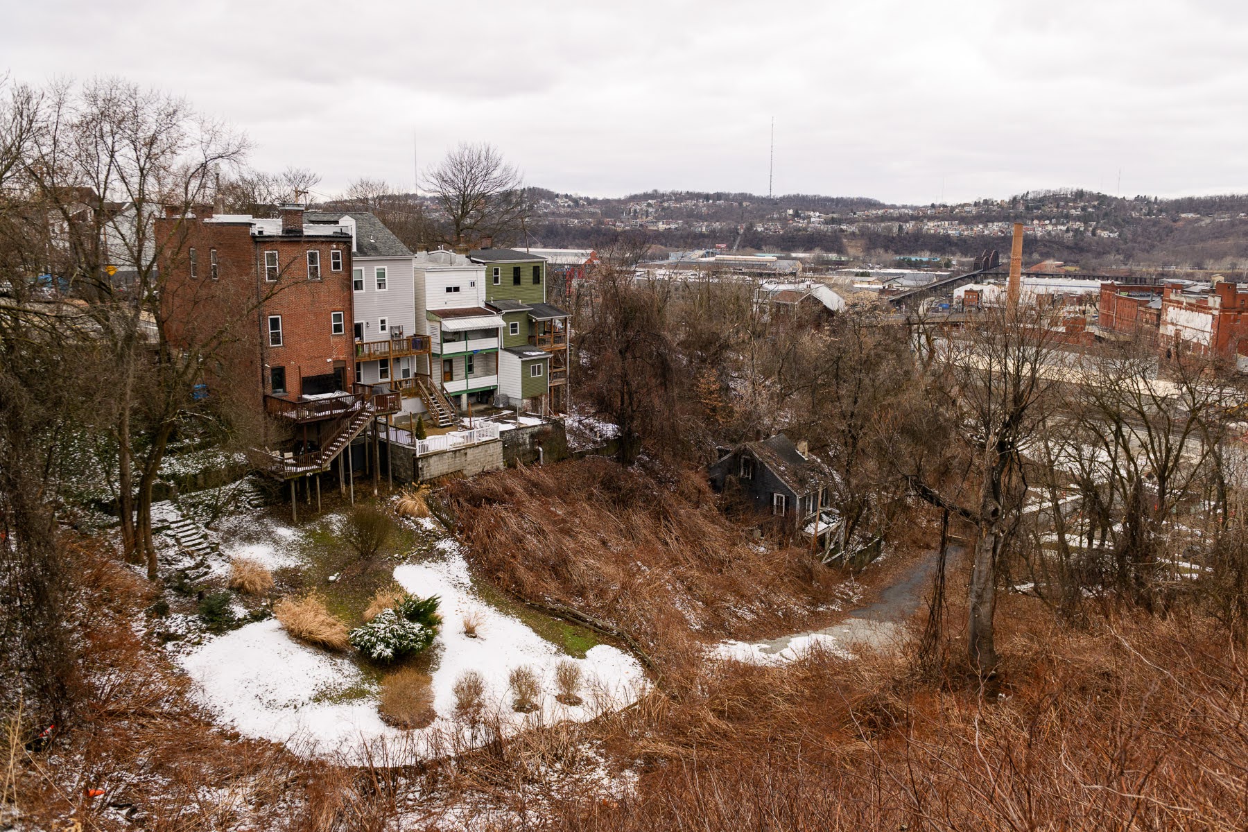 A group of houses on a hillside overlooks an industrial area with light snow on the ground and cloudy skies.