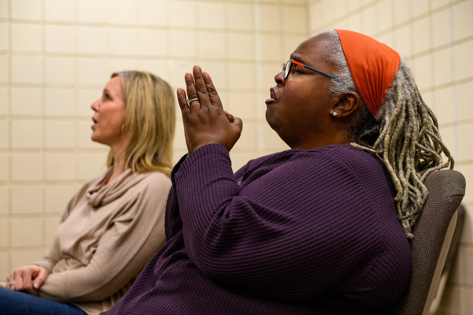 Two women sit side by side in a tiled room; one wears a purple top and orange headband with hands clasped, the other wears a beige top, both appear to be attentively listening.