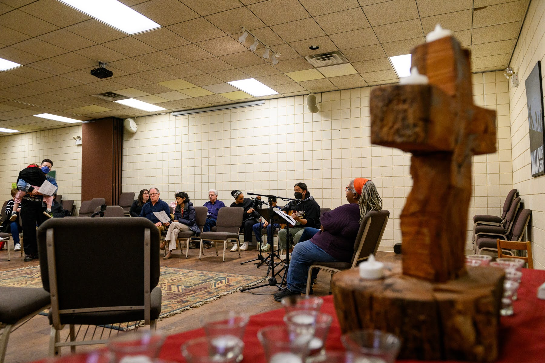 People are seated in a church or meeting room, some singing or holding papers, with a wooden cross and candles visible in the foreground.