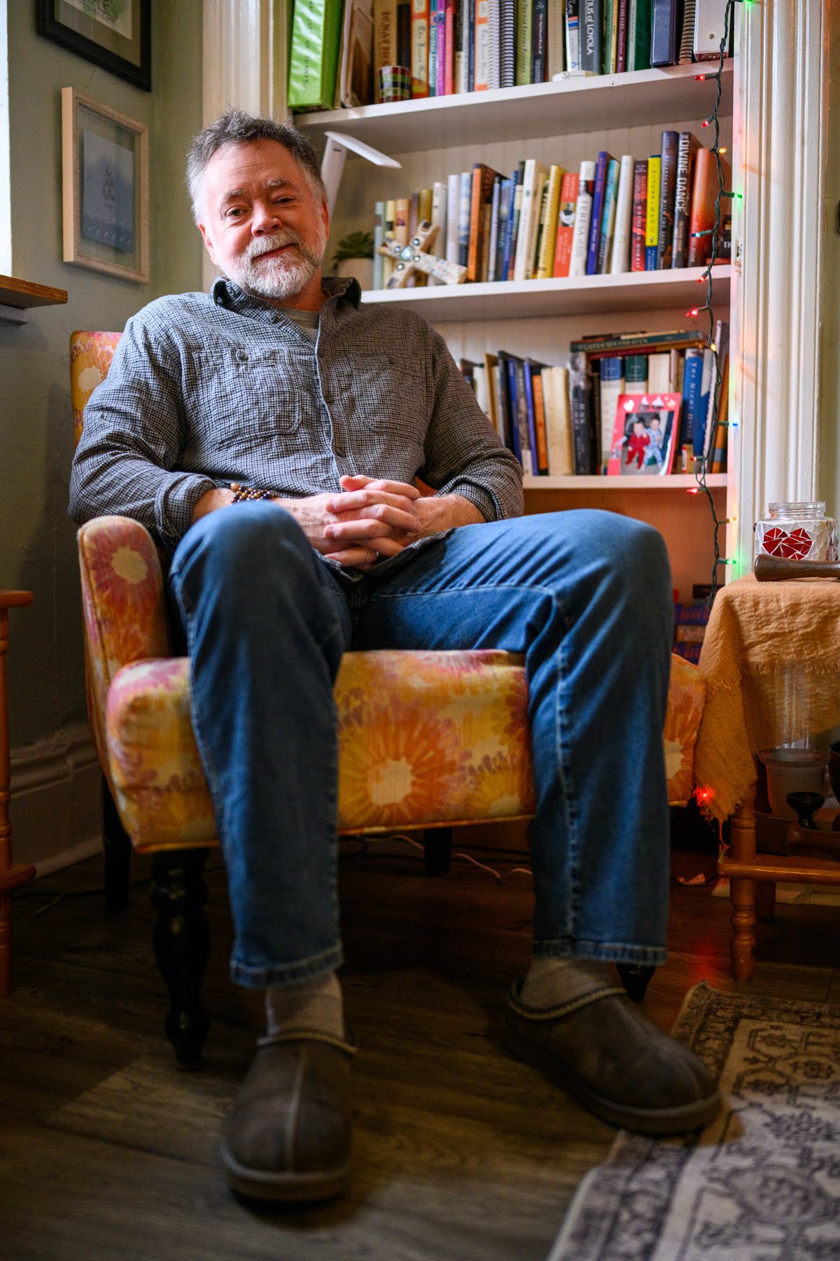 A man sits in a patterned armchair with his hands clasped, wearing a gray shirt, blue jeans, and slippers. A bookshelf and a side table are in the background.