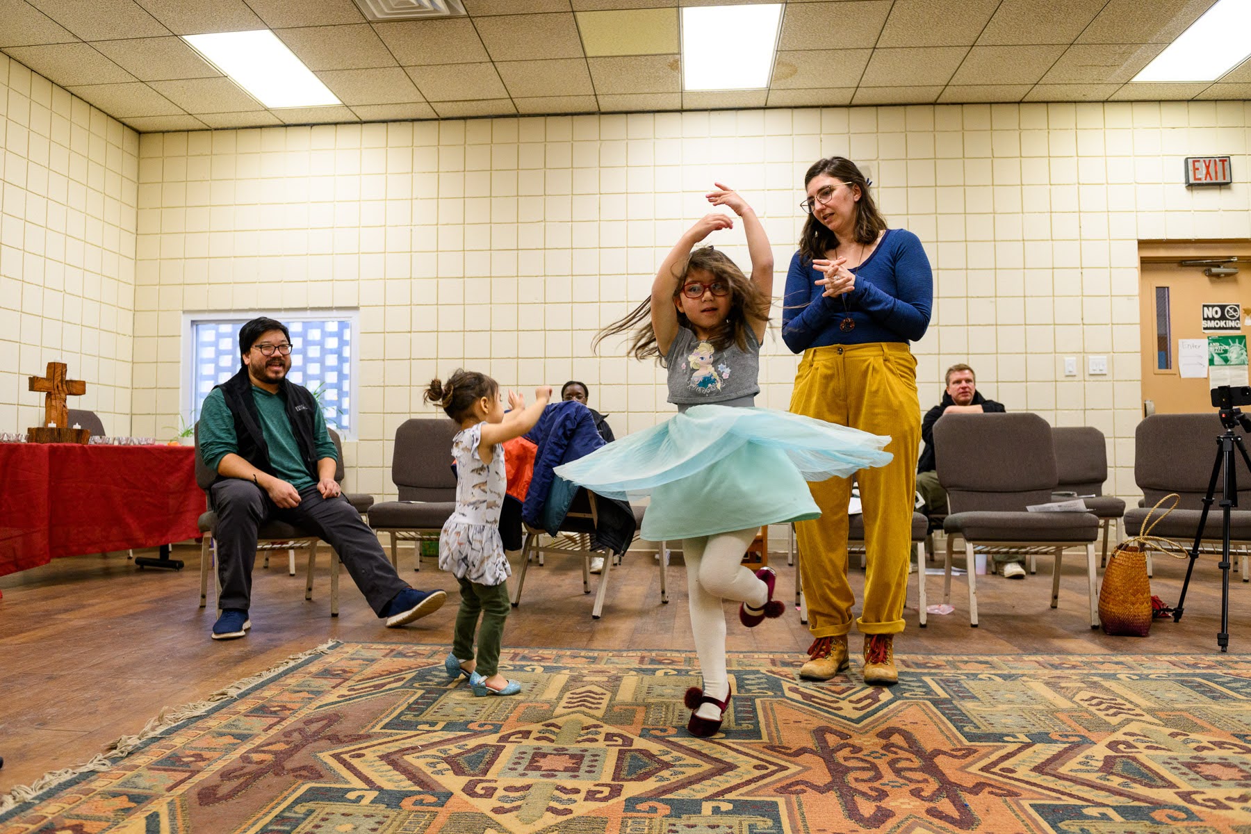Two children dance on a patterned rug while two adults watch in a room with chairs, a cross and a camera tripod in the background.