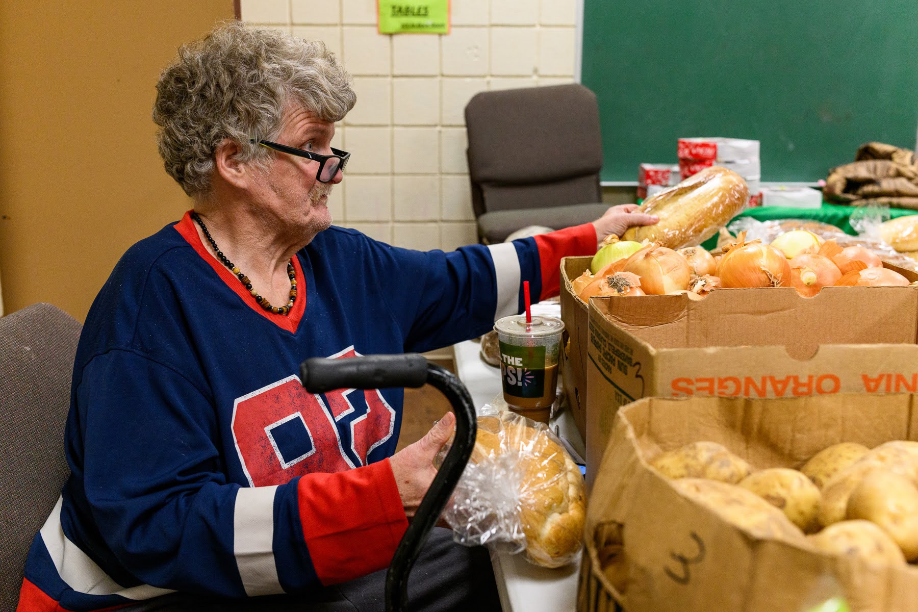 An older person in a sports jersey sits at a table sorting bread and vegetables, with boxes of onions and potatoes in front of them.