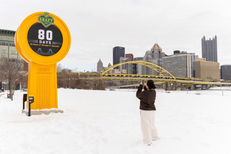 A person takes a photo of a large yellow NFL Draft countdown clock in a snowy park, with Pittsburgh's city skyline and bridges in the background.