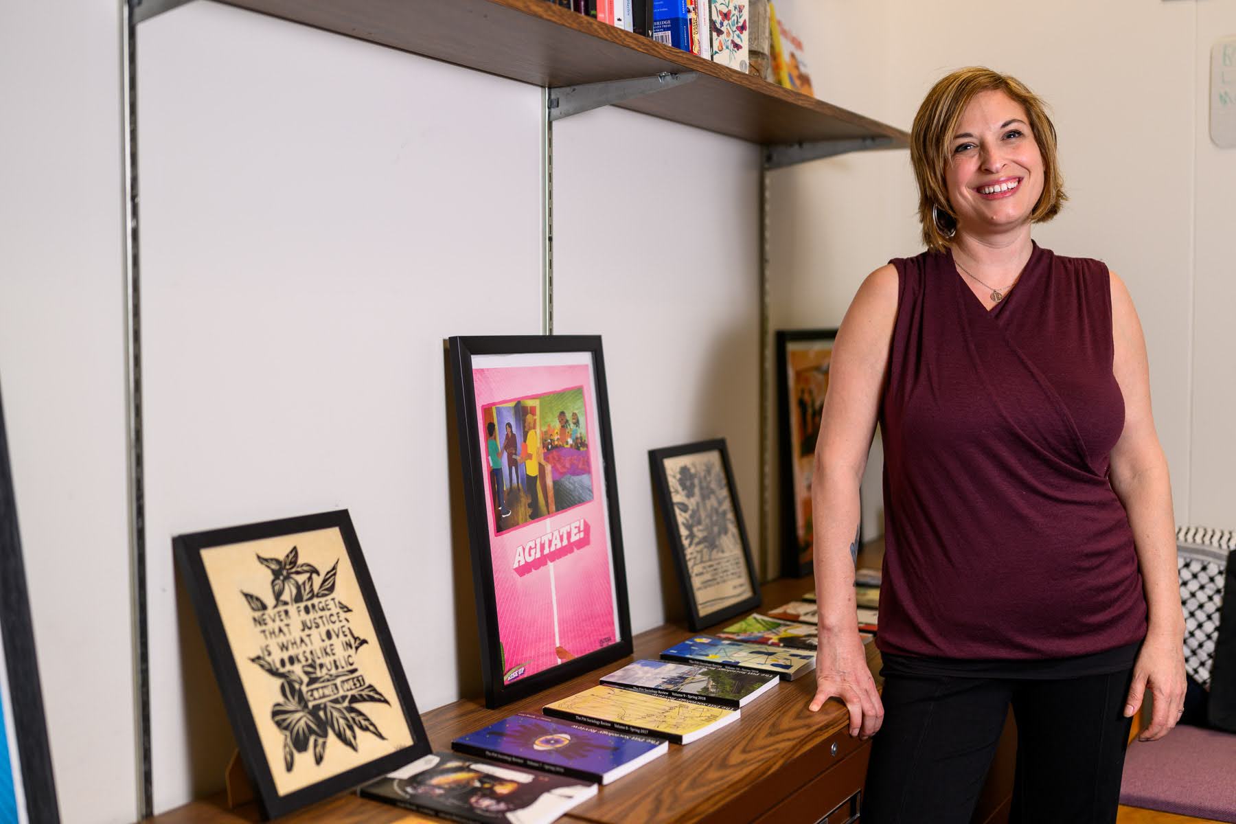 A woman stands next to a shelf displaying framed artwork, books, and prints in a well-lit room.