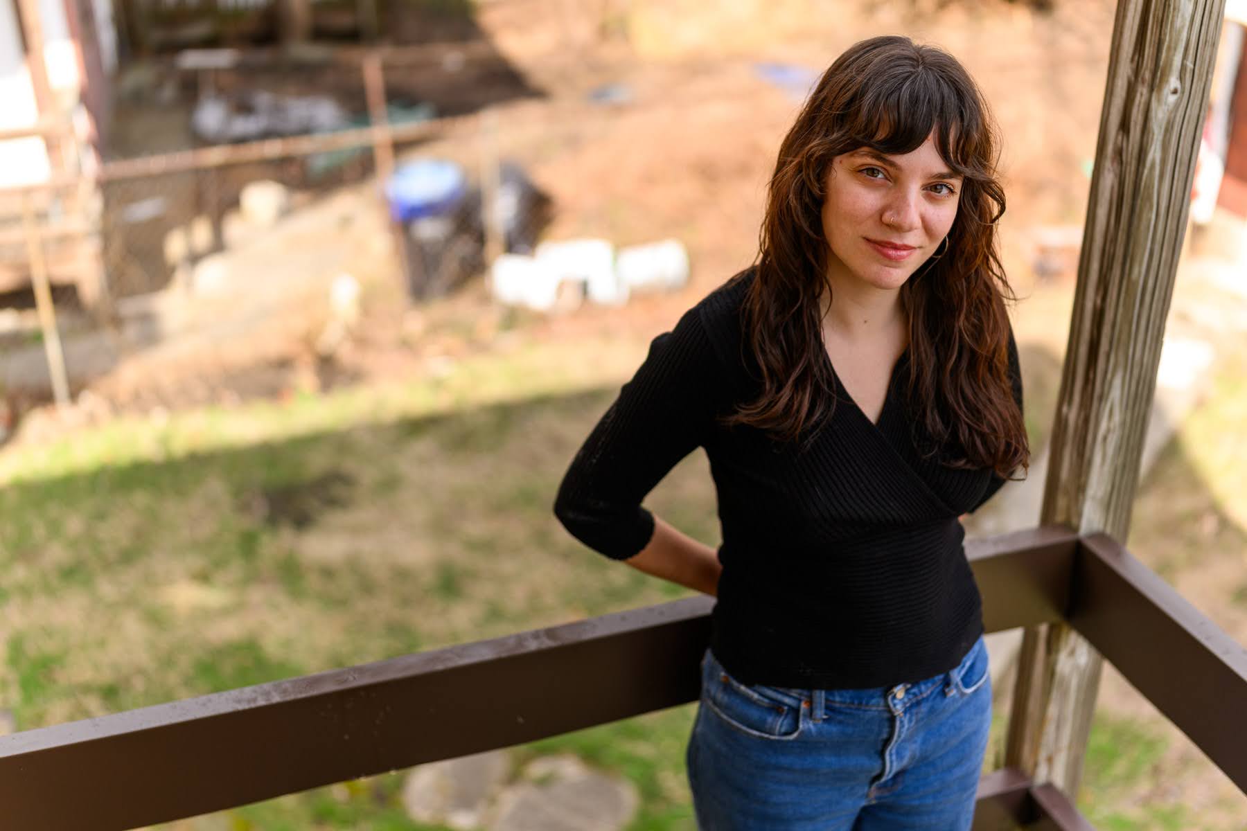 A woman with long brown hair, wearing a black top and blue jeans, stands on a porch with her hands behind her back, looking at the camera. The background shows a grassy yard.