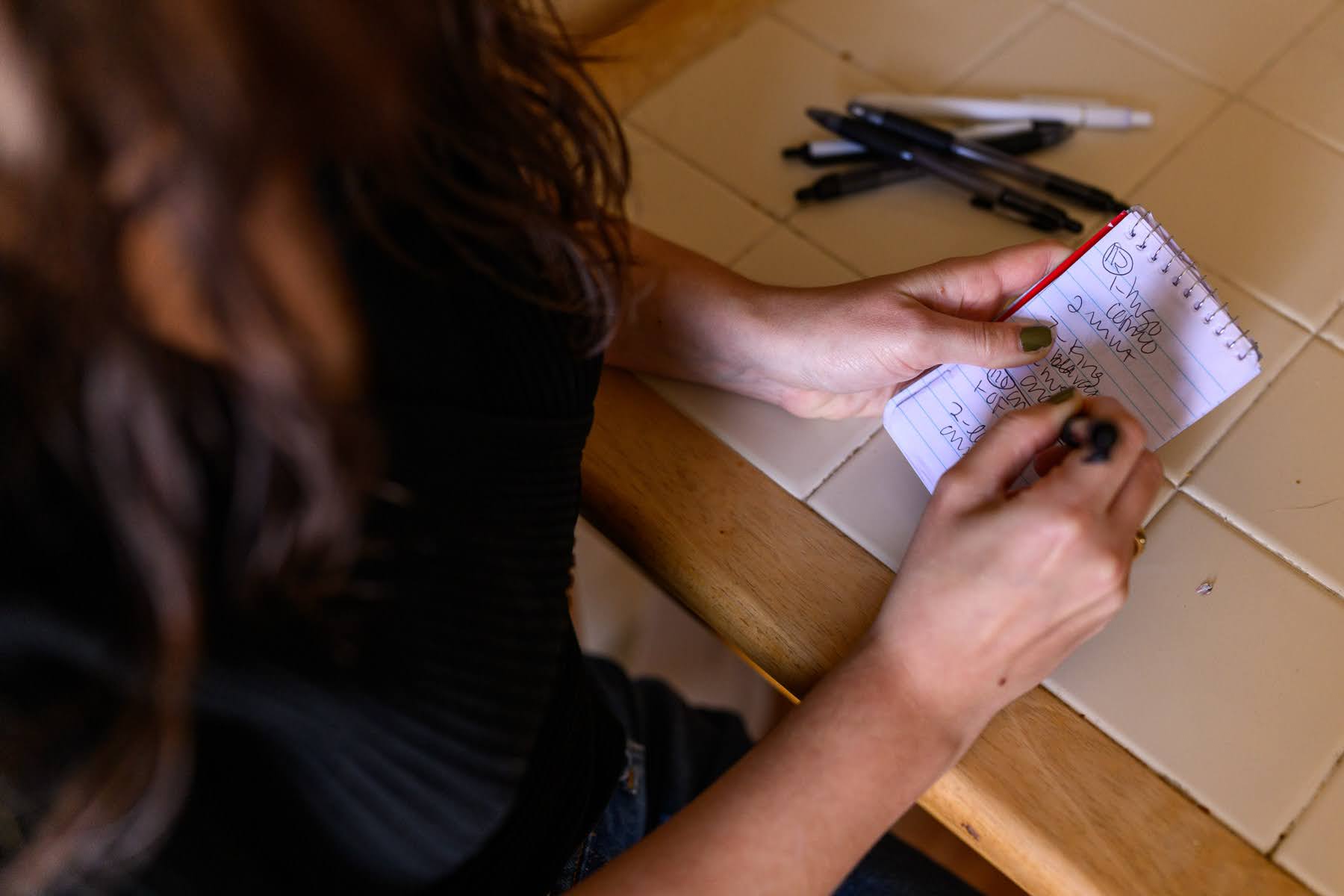 A person writes on a small notepad with multiple pens and markers on a tiled table nearby.