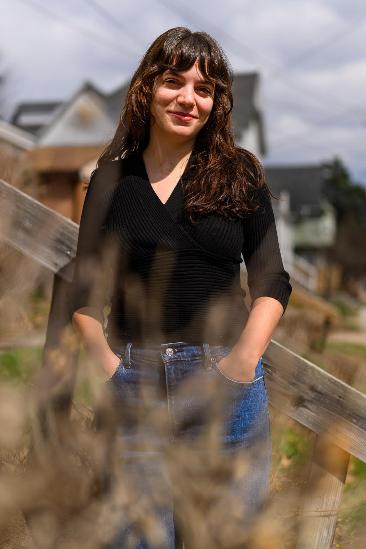 A person with long brown hair, wearing a black top and blue jeans, stands outdoors with hands in pockets, blurred plants in the foreground and houses in the background.