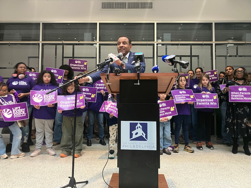 A photograph of a Black man in a suit standing behind a podium and speaking into a microphone with a group of adults and children standing in the background.