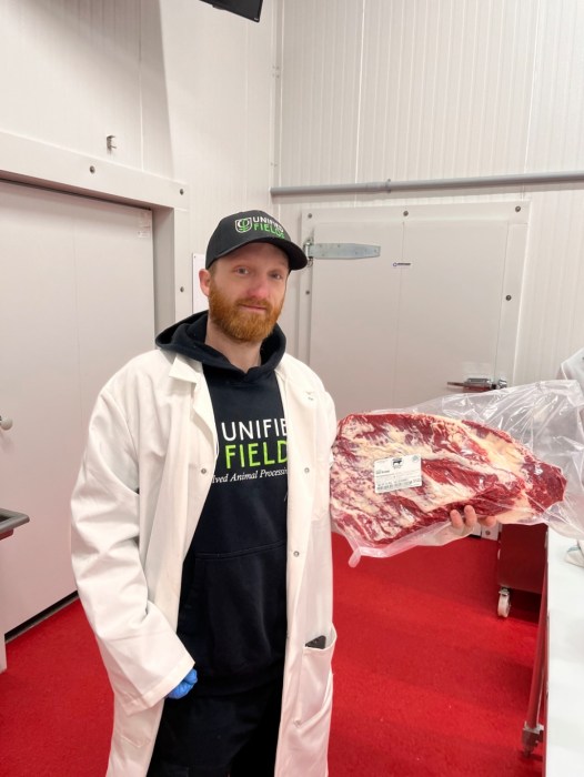 A man with a beard in a hat standing next to a large cut of meat
