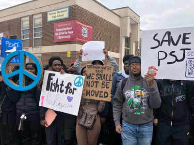 A photograph of student protestors holding signs standing outside of a large brick school building.