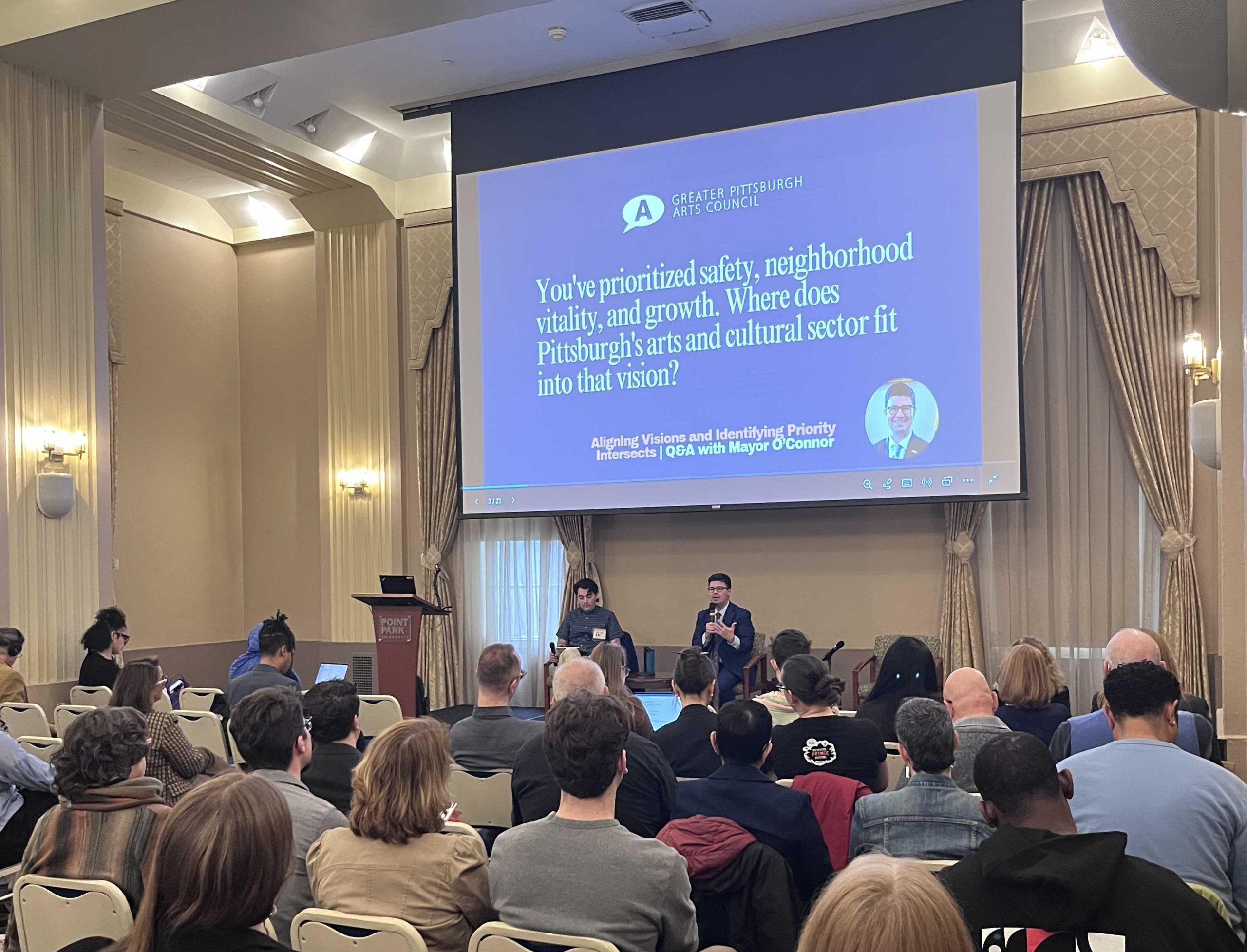 A speaker and moderator engage in a Q&A session on stage with an audience, discussing Pittsburgh’s arts and cultural sector, as shown on a projection screen behind them.