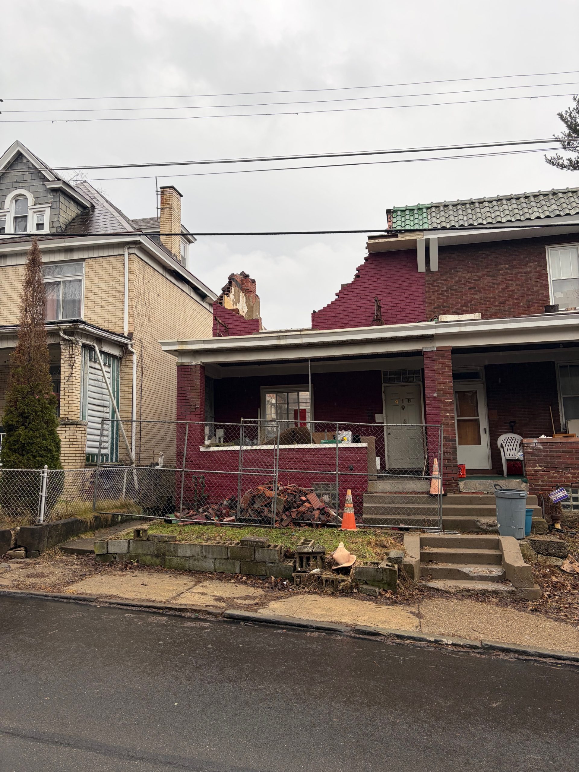 A partially collapsed red brick house with a damaged roof and debris on the porch, next to an intact neighboring home on a cloudy day.