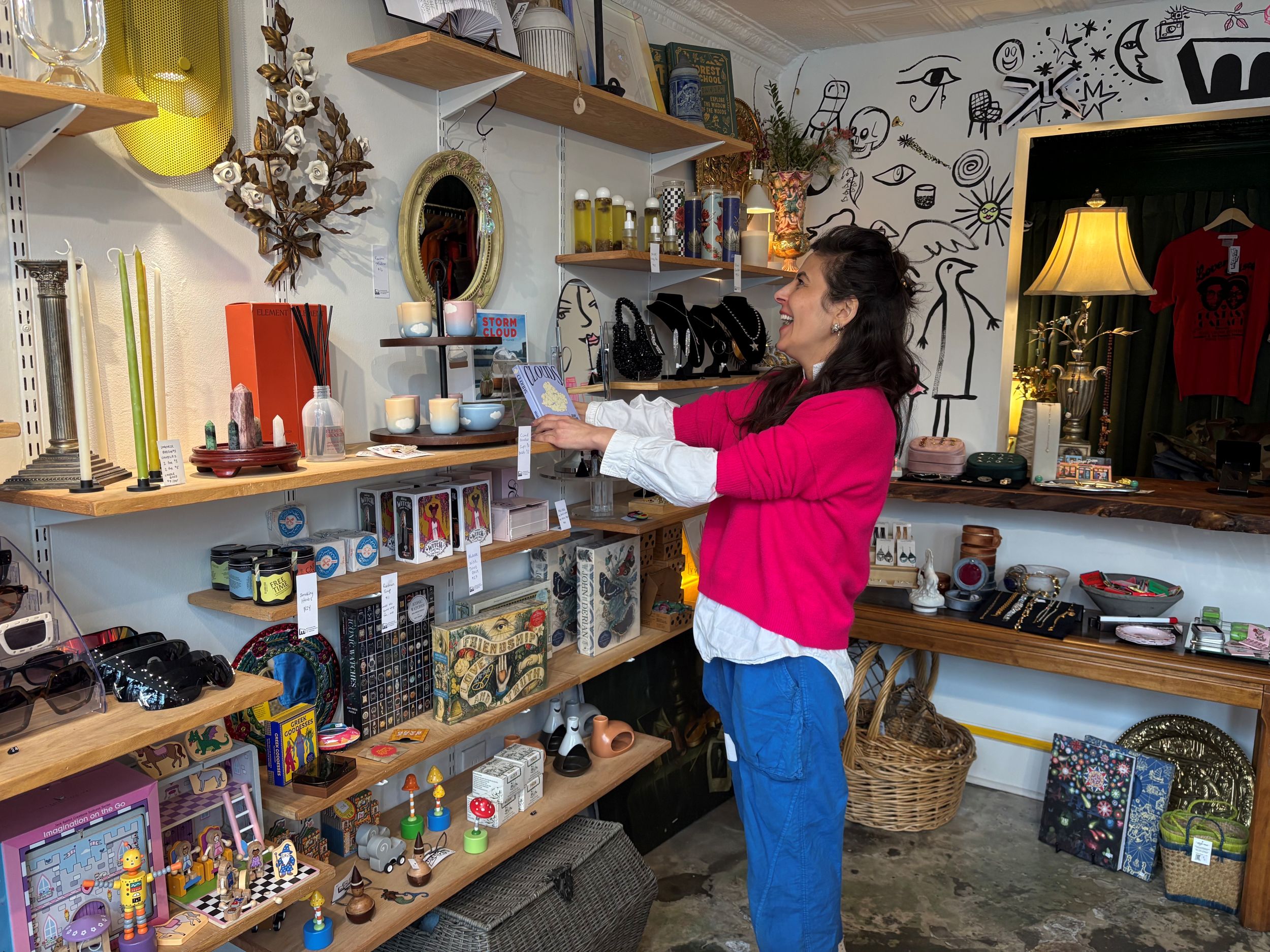 A woman in a pink sweater and blue pants smiles while browsing items on a shelf in a small, eclectic shop filled with various decor and gifts.
