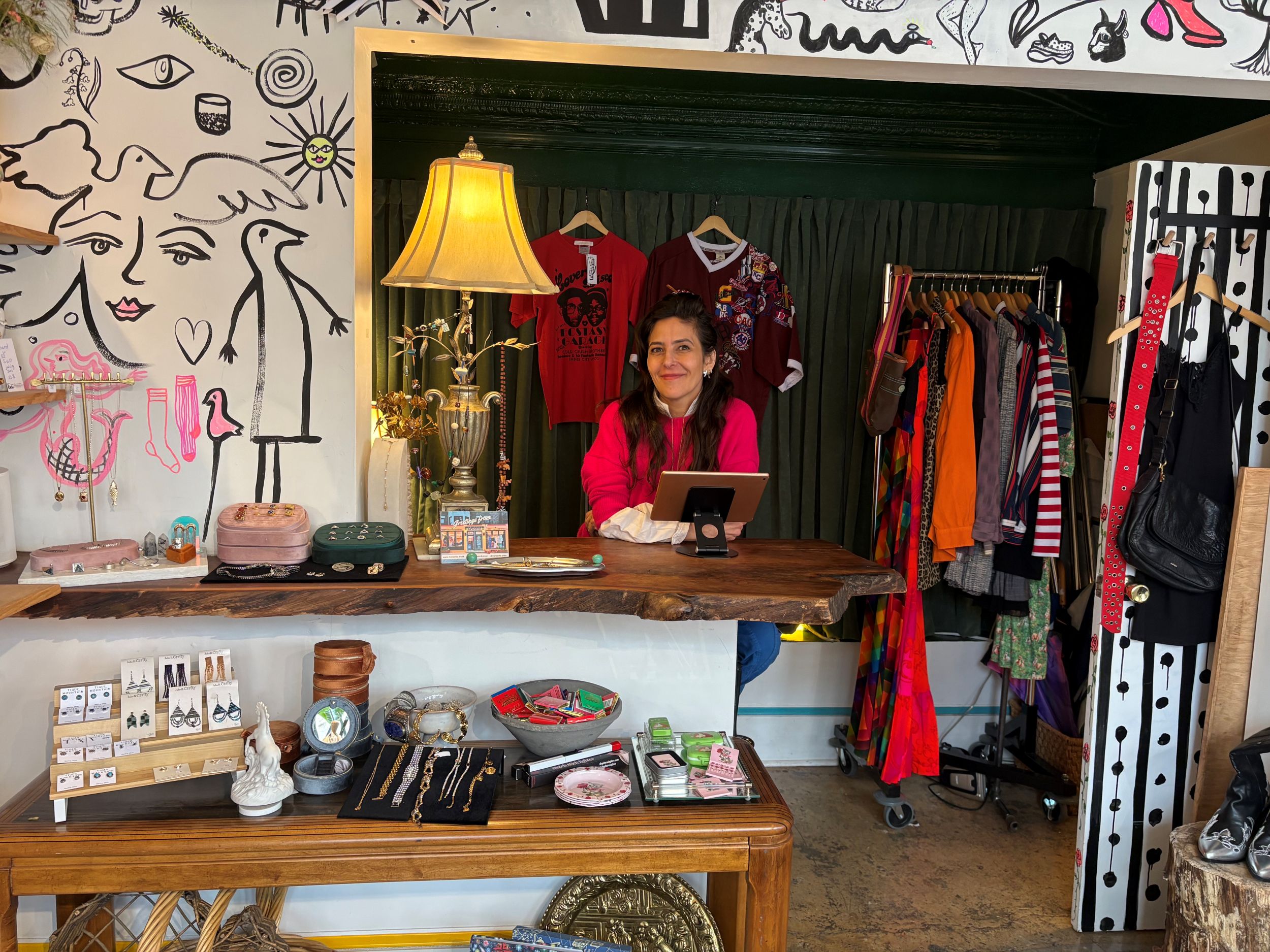 A woman sits behind a wooden counter in a boutique shop with clothing, jewelry, and decor items on display. Hand-drawn designs cover the walls behind her.