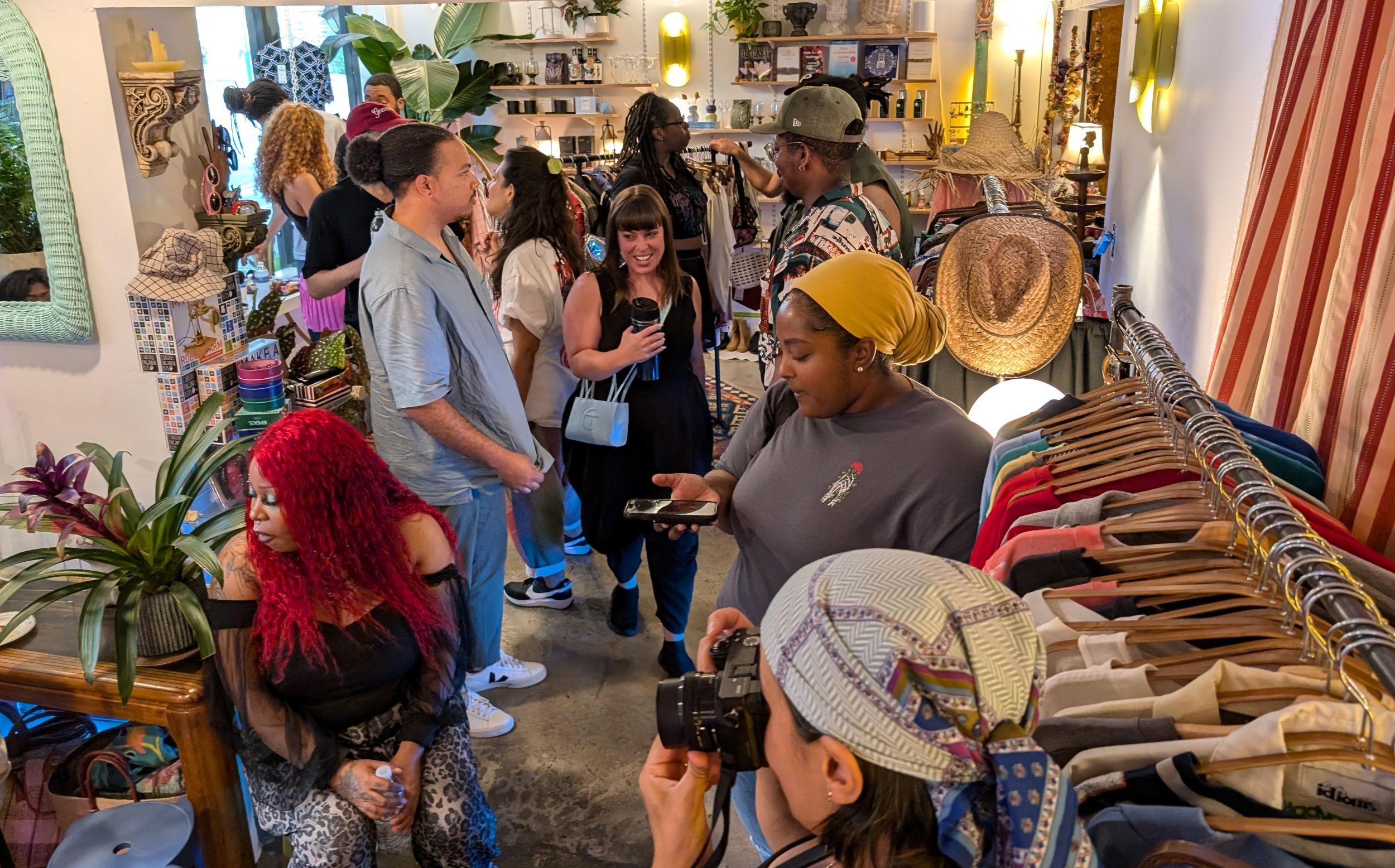 A group of people socialize and browse clothing racks inside a brightly lit boutique with plants and eclectic decor.