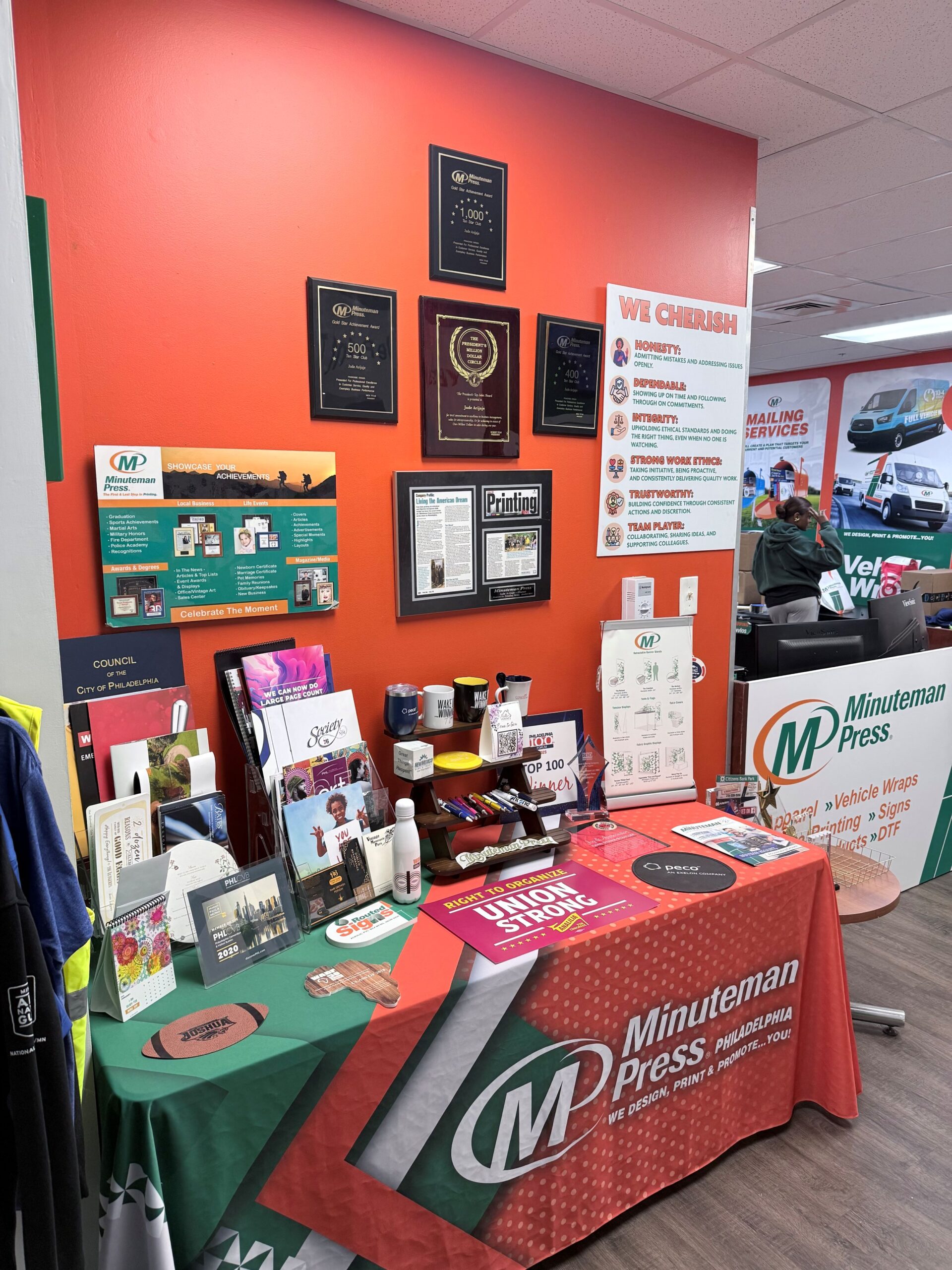A display table with Minuteman Press promotional items and brochures is set up against an orange wall featuring awards and certificates in a print shop.