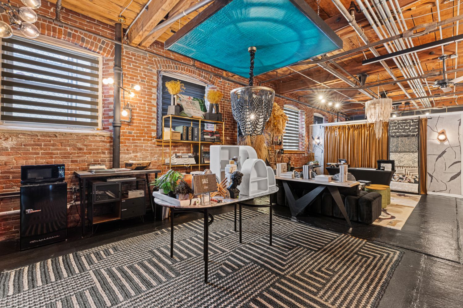Modern office space with exposed brick walls, wooden ceiling, and industrial pipes, featuring a meeting table, lounge area, shelves, and decorative lighting fixtures.