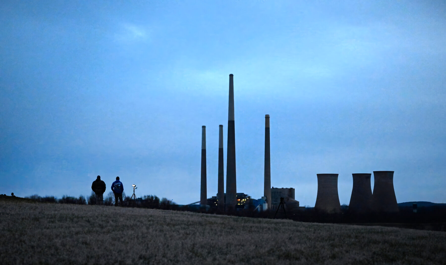 Two people walk across a grassy field at dusk with several tall smokestacks and cooling towers of a power plant in the background.